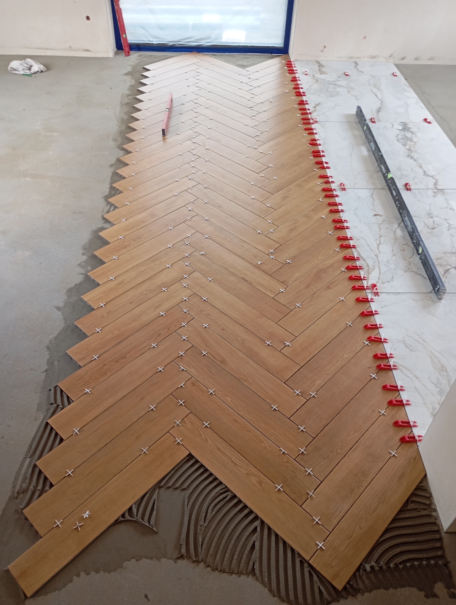 Układanie drewnopodobnych płytek podłogowych w jodełkę, z użyciem krzyżyków dystansowych i czerwonych klipsów poziomujących, widoczny fragment ściany i okna.