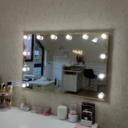 Toaletka z podświetlanym lustrem w złotym obramowaniu, odbijająca wnętrze pokoju z krzesłem barowym i neonem. Na blacie kosmetyki i akcesoria.