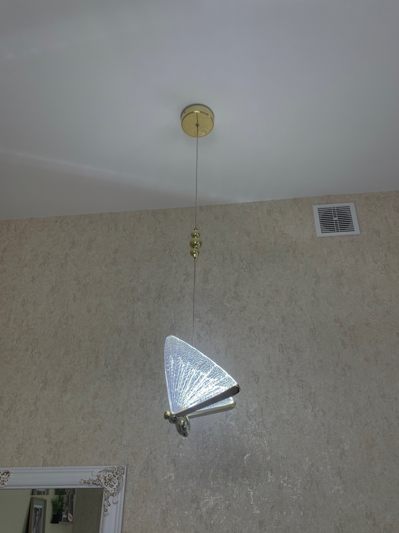 Zawieszona lampa w kształcie motyla z dekoracyjnymi elementami, złota podsufitka, na tle ściany z beżową tapetą i białego sufitu.