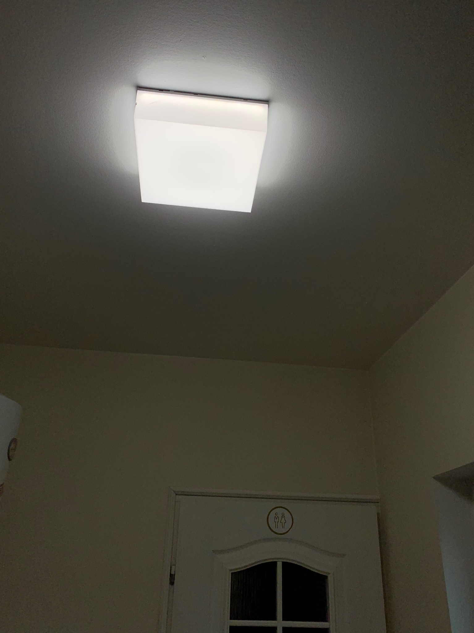Zamontowana, kwadratowa lampa sufitowa LED, oświetlająca pomieszczenie z białą ścianą i drzwiami z symbolem toalety. Minimalistyczny design, ciepłe światło.