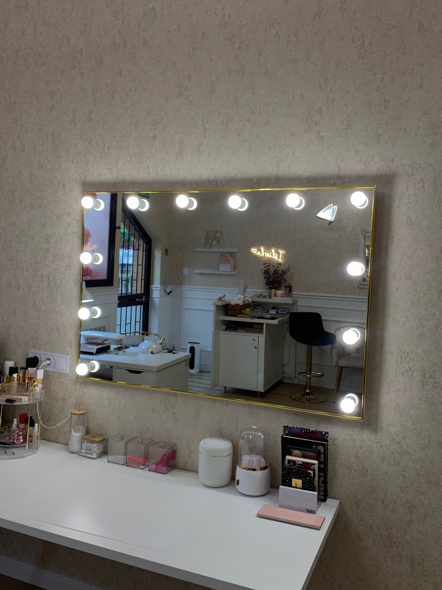 Toaletka z podświetlanym lustrem w złotym obramowaniu, odbijająca wnętrze pokoju z krzesłem barowym i neonem. Na blacie kosmetyki i akcesoria.