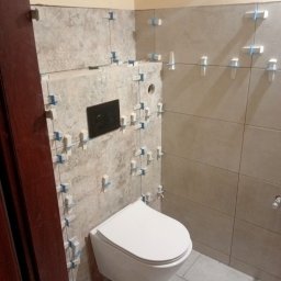 Pawel Gasior - Świeżo ułożone, beżowe płytki w łazience, z widocznymi krzyżykami dystansowymi, wisząca toaleta z czarnym przyciskiem spłukującym, szare płytki podłogowe, fragment brązowej ościeżnicy drzwi.