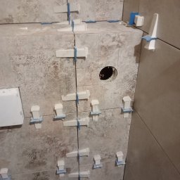 Pawel Gasior - Świeżo ułożone płytki ścienne w łazience, z użyciem białych klinów i niebieskich krzyżyków dystansowych do utrzymania równych spoin; widoczne wycięcie pod gniazdko elektryczne.