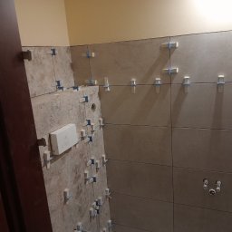 Pawel Gasior - Układanie beżowych płytek w łazience, widoczne krzyżyki dystansowe i wystające rury instalacji wodnej, ściana pomalowana na żółto, fragment ościeżnicy drzwi.