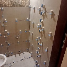 Pawel Gasior - Proces układania beżowych płytek w łazience z wykorzystaniem klipsów poziomujących i krzyżyków dystansowych, widoczne surowe ściany i instalacja wodna.