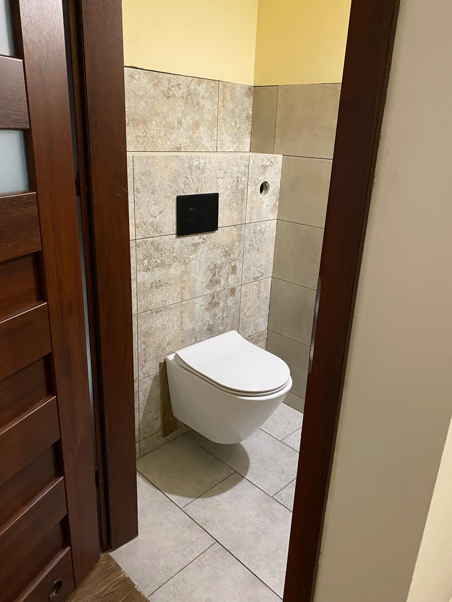 Zamontowana podwieszana toaleta z czarnym panelem sterującym na ścianie wyłożonej beżowymi płytkami imitującymi kamień, widoczna zza otwartych, ciemnobrązowych drzwi w łazience z jasnymi płytkami...