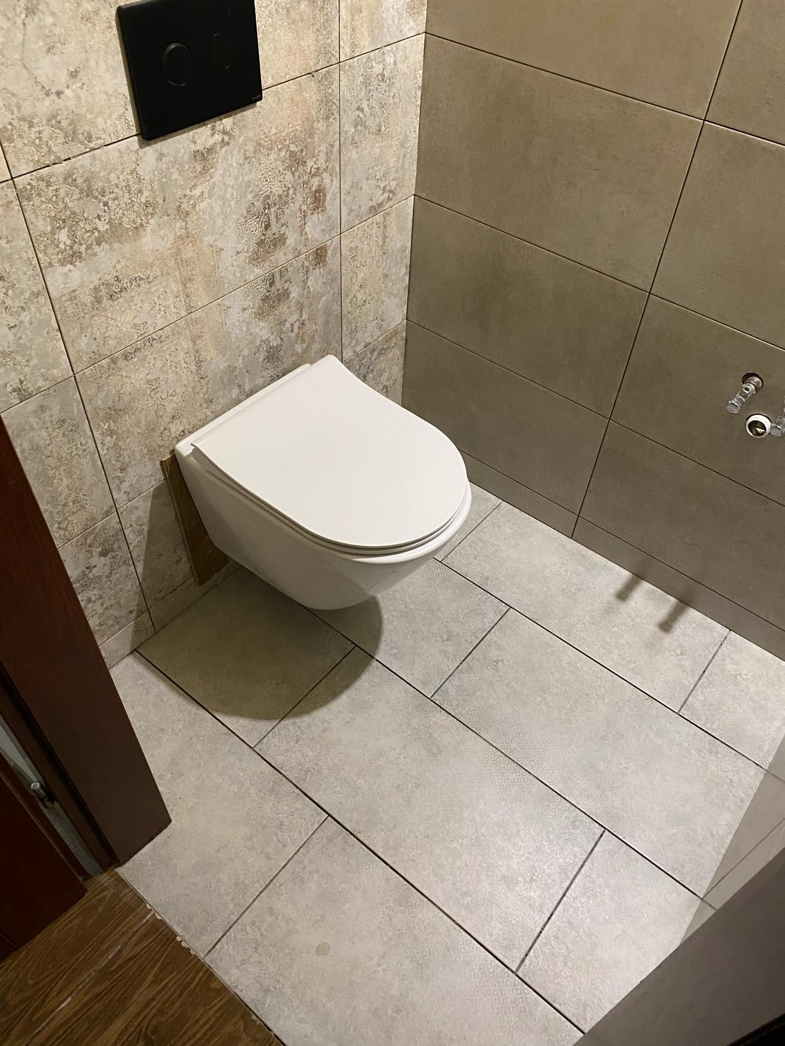 Wisząca miska WC z podtynkową spłuczką na tle beżowych płytek w łazience po remoncie, widoczny fragment drewnianej futryny drzwiowej.