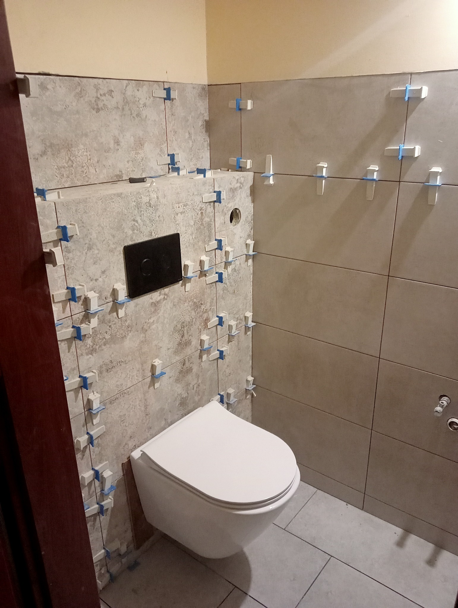 Świeżo ułożone płytki ścienne w łazience, z białymi krzyżykami dystansowymi i czarnym panelem sterowania spłuczką nad podwieszaną toaletą.