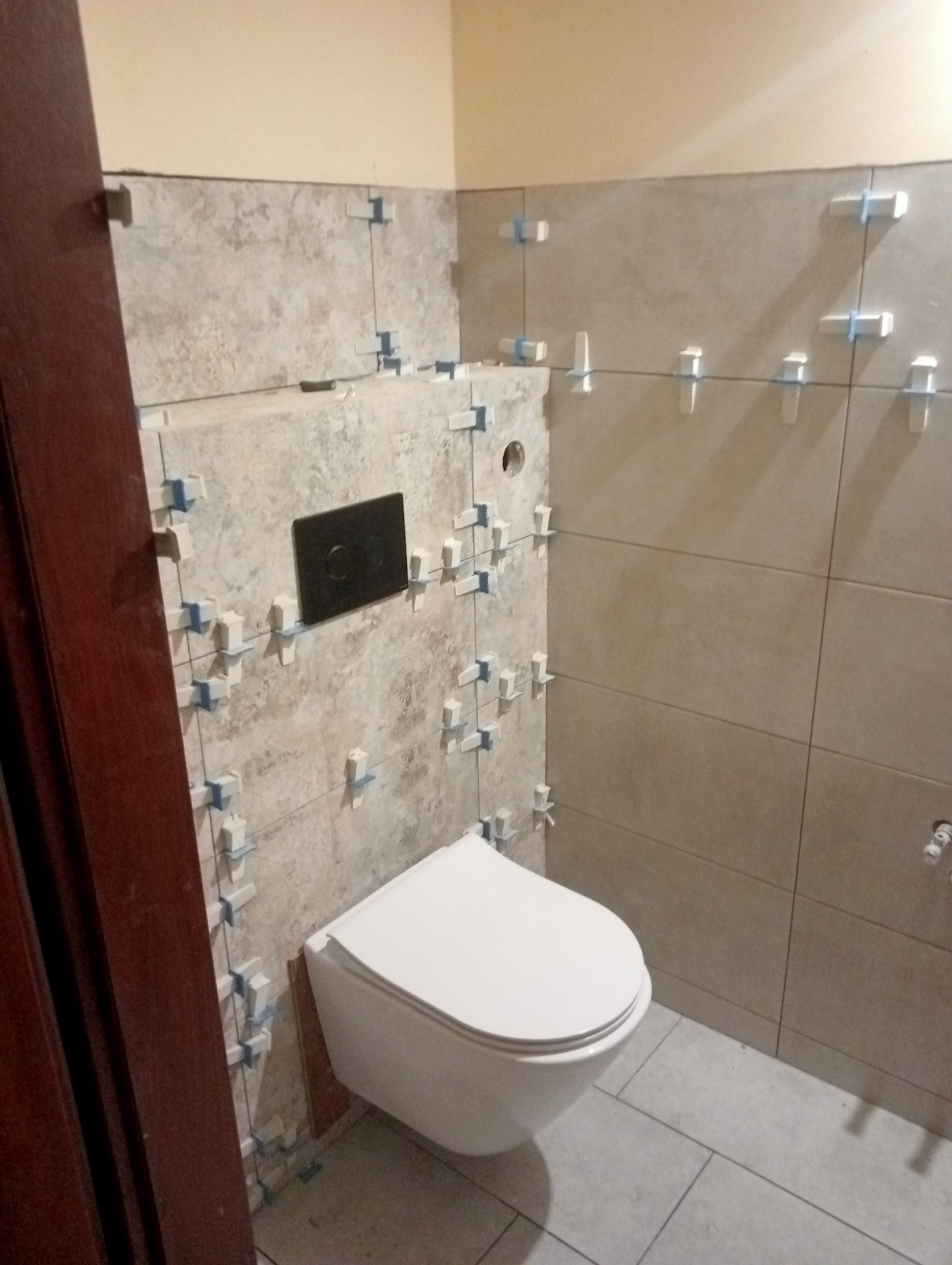 Świeżo ułożone, beżowe płytki w łazience, z widocznymi krzyżykami dystansowymi, wisząca toaleta z czarnym przyciskiem spłukującym, szare płytki podłogowe, fragment brązowej ościeżnicy drzwi.