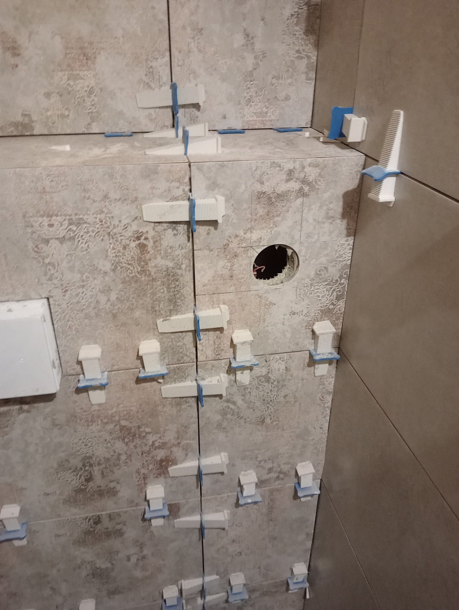 Świeżo ułożone płytki ścienne w łazience, z użyciem białych klinów i niebieskich krzyżyków dystansowych do utrzymania równych spoin; widoczne wycięcie pod gniazdko elektryczne.