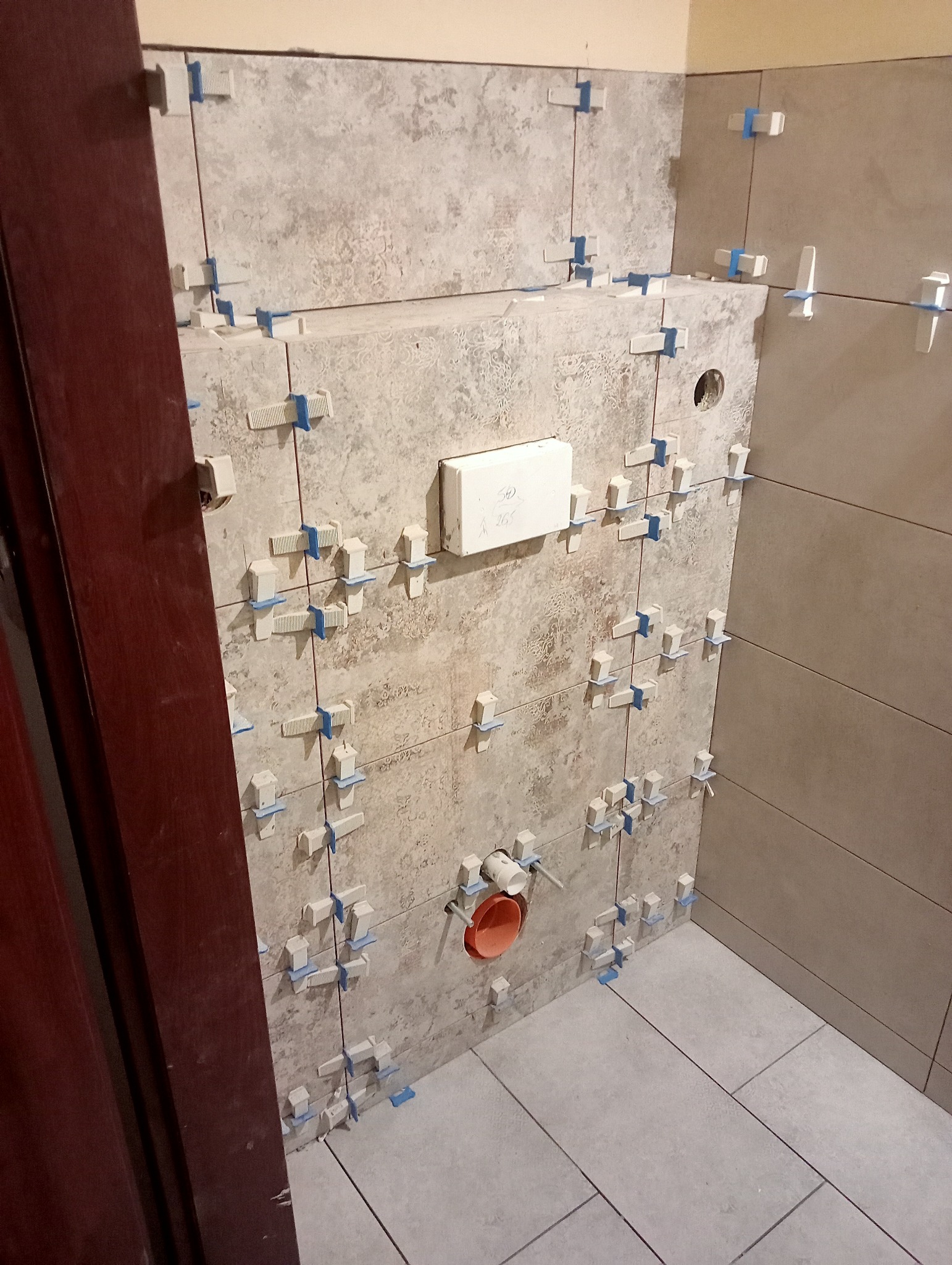 Ściana łazienki w trakcie układania płytek ceramicznych z użyciem krzyżyków dystansowych, widoczny otwór na instalację sanitarną i częściowo ułożona podłoga.