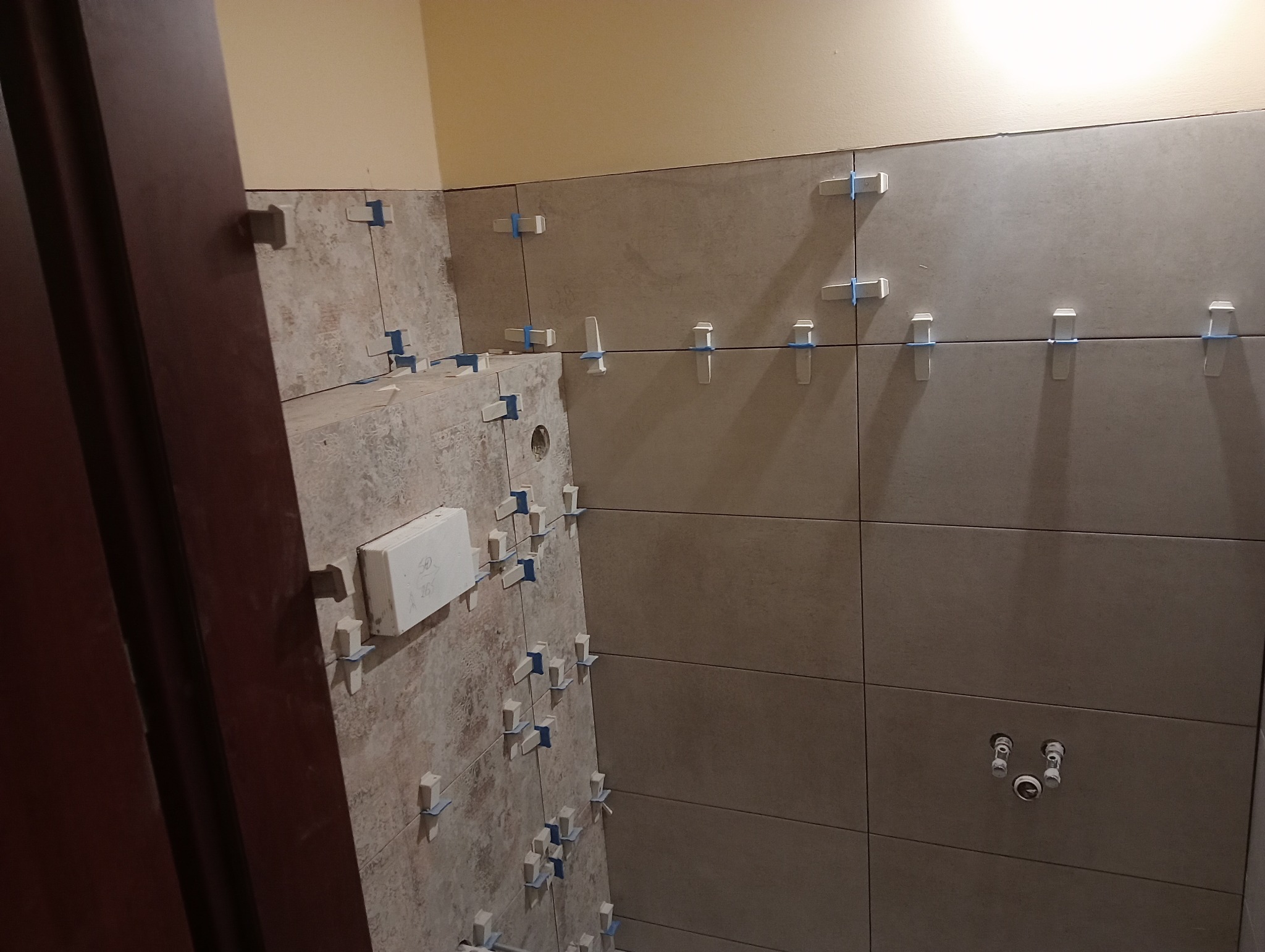 Układanie beżowych płytek w łazience, widoczne krzyżyki dystansowe i wystające rury instalacji wodnej, ściana pomalowana na żółto, fragment ościeżnicy drzwi.