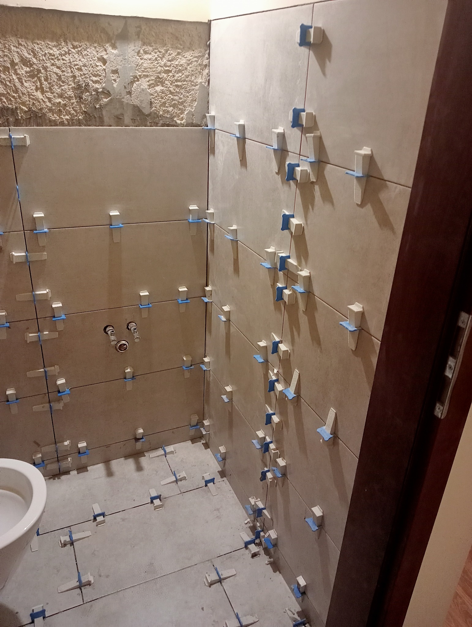 Proces układania beżowych płytek w łazience z wykorzystaniem klipsów poziomujących i krzyżyków dystansowych, widoczne surowe ściany i instalacja wodna.