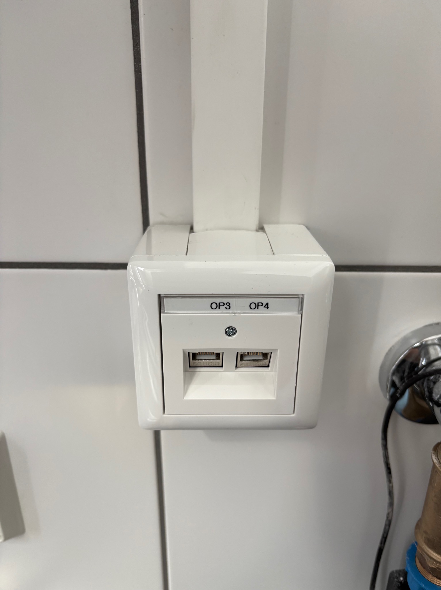 Białe gniazdo sieciowe (OP3, OP4) zamontowane na kafelkowanej ścianie, z widoczną rurką instalacyjną i fragmentem instalacji wodnej po prawej stronie.
