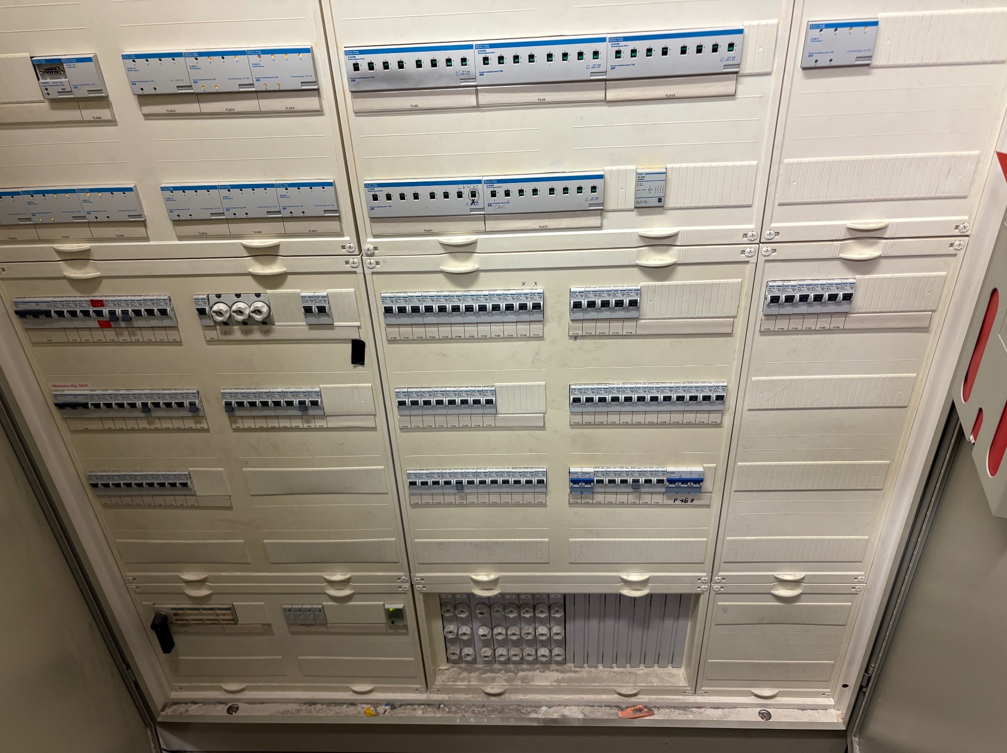 Rozbudowana, biała skrzynka elektryczna z licznymi bezpiecznikami, wyłącznikami i modułami, widoczna pod kątem z dołu, ukazująca złożoność instalacji.