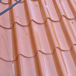 GREEN MASTER MYKHAILO BURDOVSKYI - Błyszczące, mokre dachówki z blachy trapezowej w kolorze ceglastym, widziane z góry, z fragmentem buta robotnika w lewym dolnym rogu i wężem ogrodowym na górze.