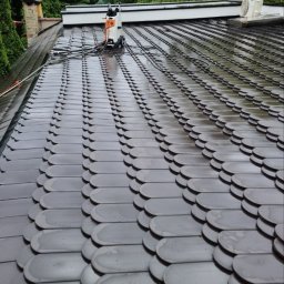 GREEN MASTER MYKHAILO BURDOVSKYI - Czyszczenie dachu z dachówki bitumicznej za pomocą myjki ciśnieniowej, widoczne ślady czyszczenia, w tle drzewa i komin.