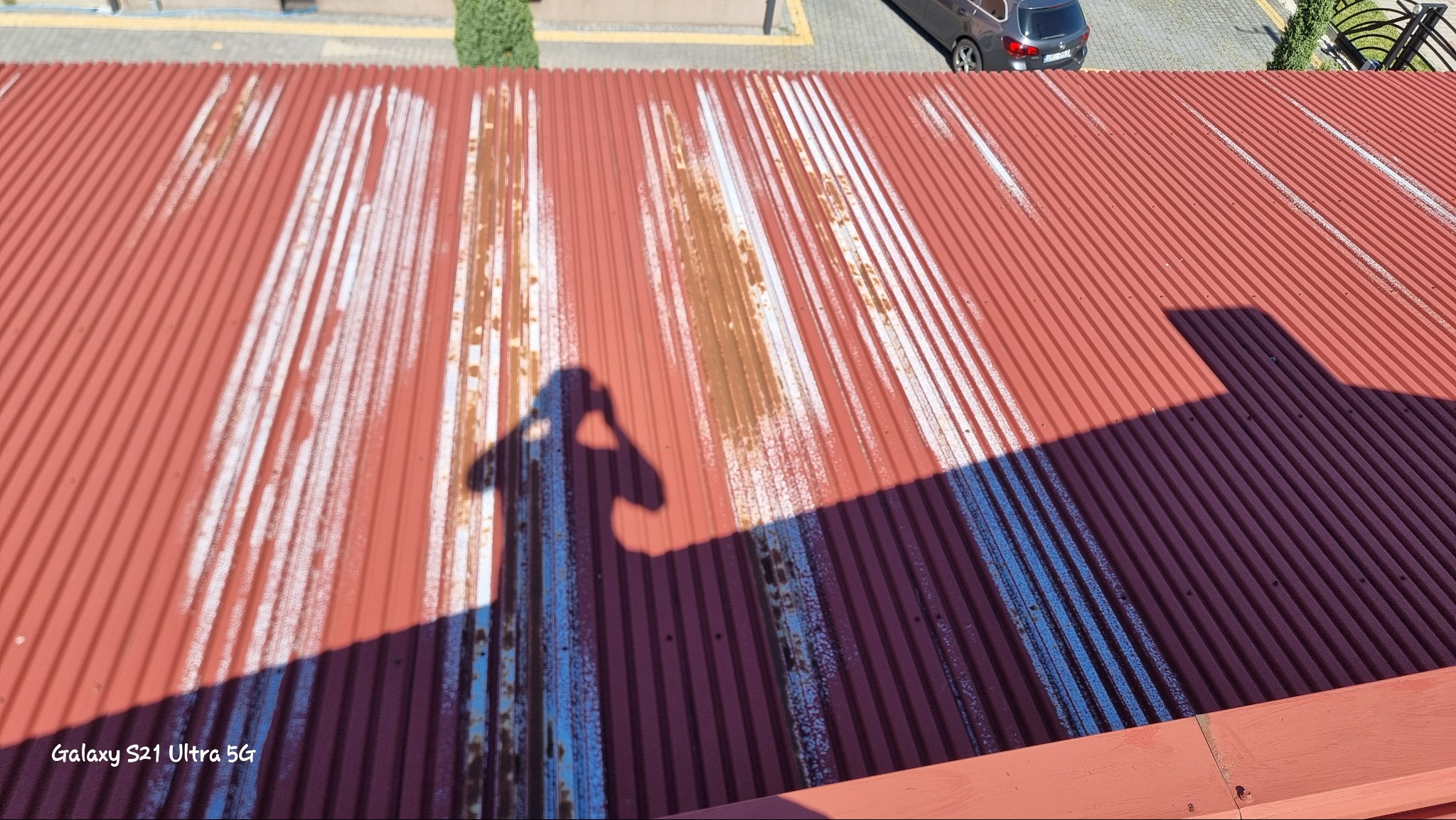 Czerwona, pofałdowana blacha z widocznymi śladami korozji i resztkami białej farby, cień fotografa w lewym centrum kadru, fragment samochodu i zieleni w tle, widok z góry.