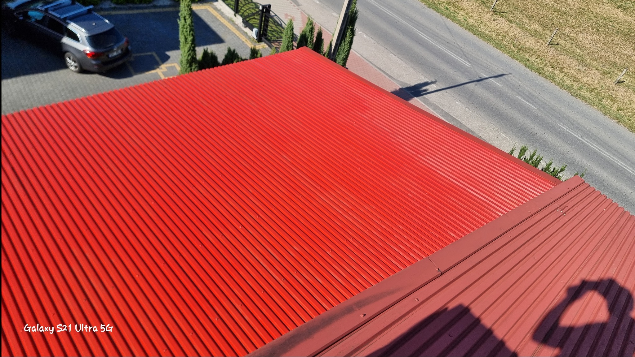 Czerwony blaszany dach z widoczną fakturą, sfotografowany z góry, z fragmentem ulicy i samochodu w tle.