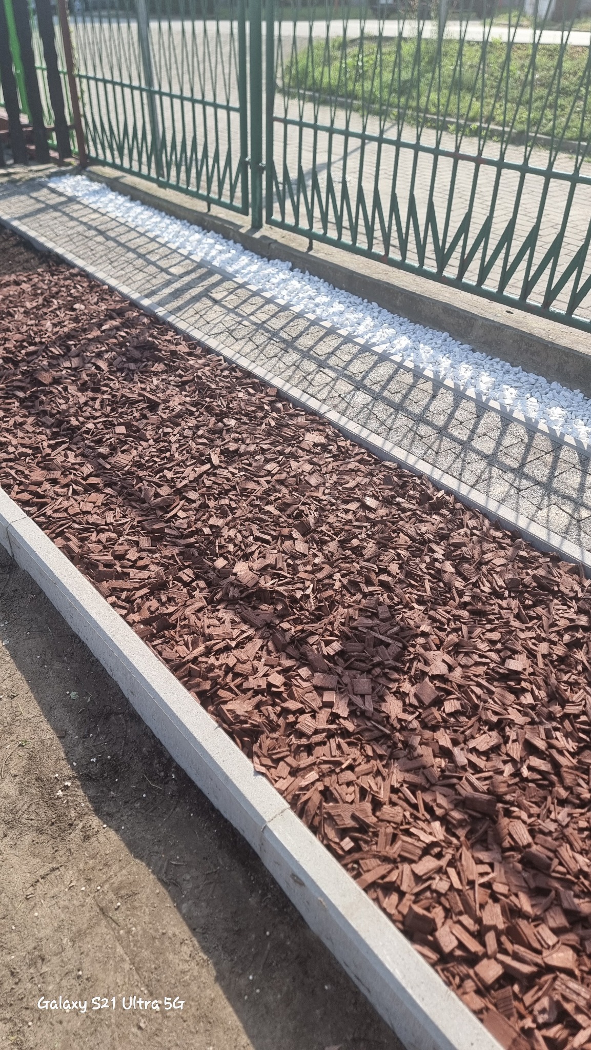Krawężnik oddzielający rabatę z kory drzewnej od białych kamyków przy metalowym ogrodzeniu.