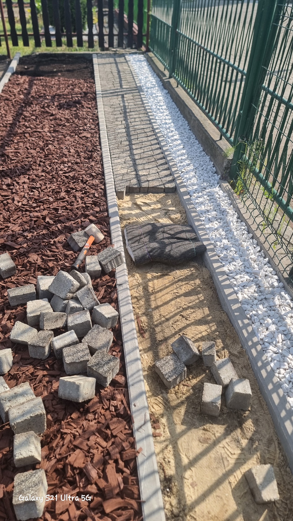 Fragment układanej ścieżki z kostki brukowej; po lewej stronie kora drzewna, po prawej białe kamienie ozdobne, w tle zielone ogrodzenie i młotek brukarski.