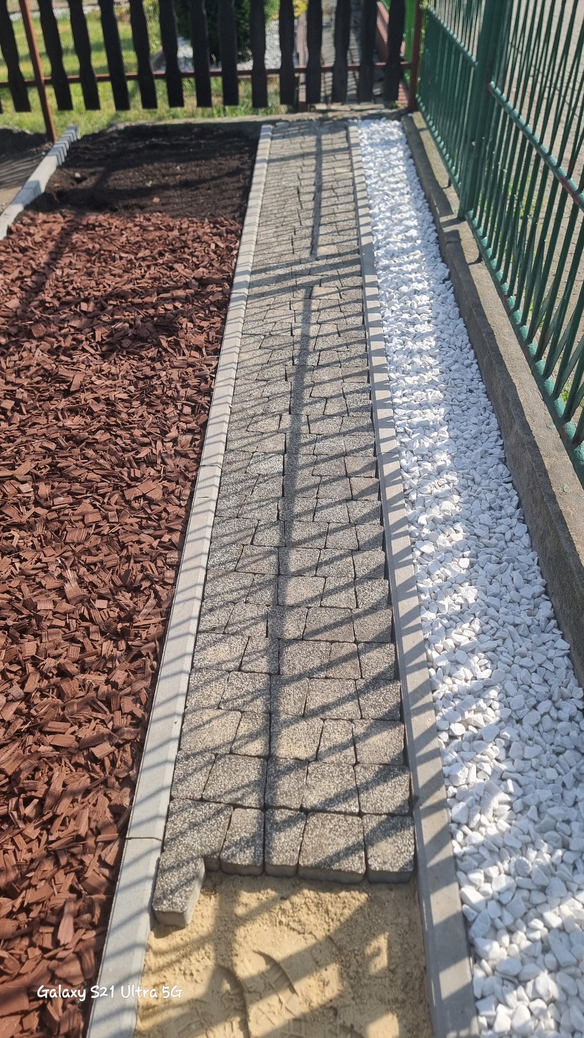 Ścieżka z szarej kostki brukowej w trakcie układania, obok kory drzewnej i białych kamyków, widoczne odciski butów w piasku.