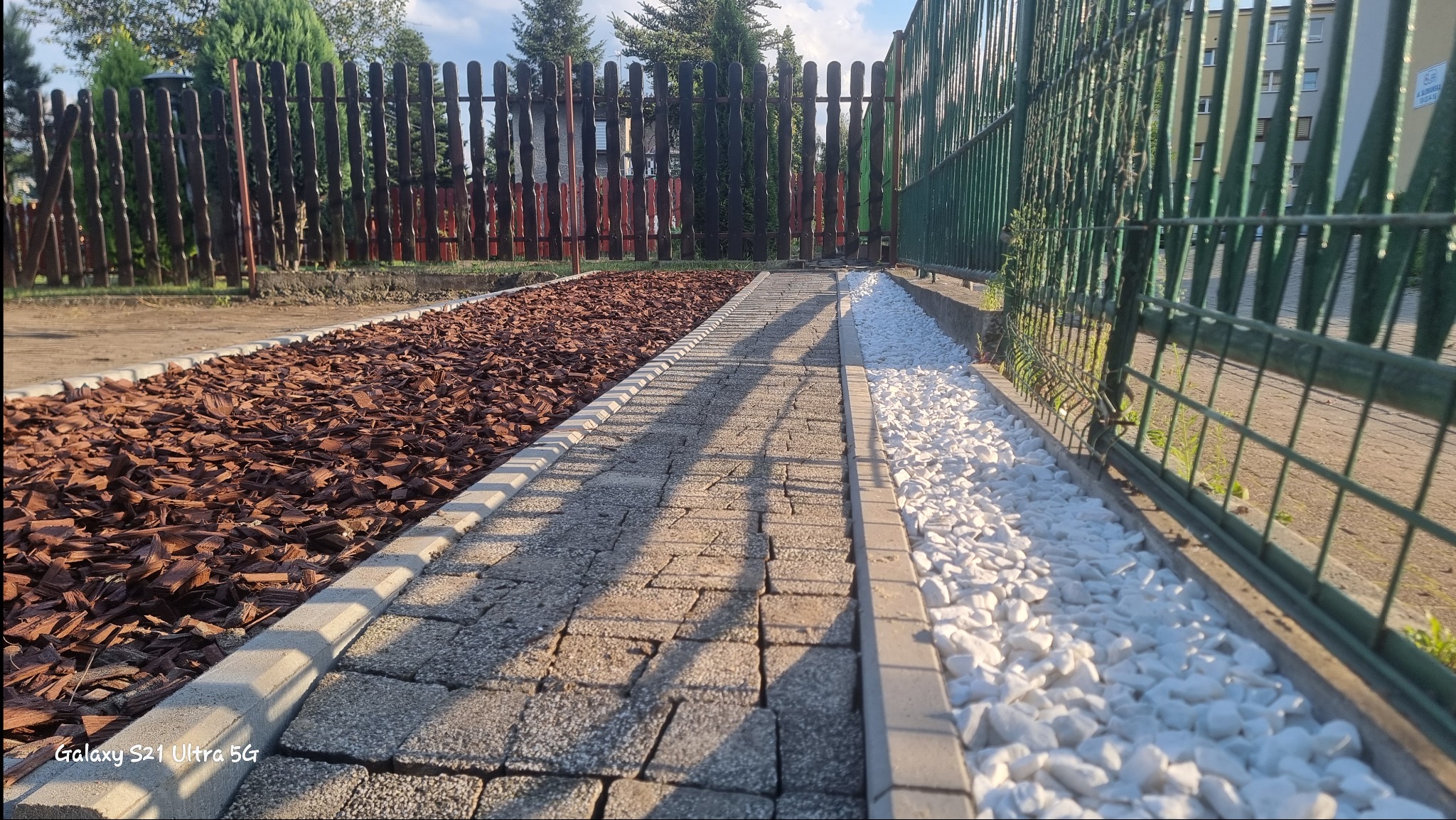 Ścieżka z kostki brukowej obramowana brązowymi zrębkami kory po lewej i białymi kamieniami po prawej stronie, widok wzdłuż ogrodzenia z siatki i drewnianego płotu.