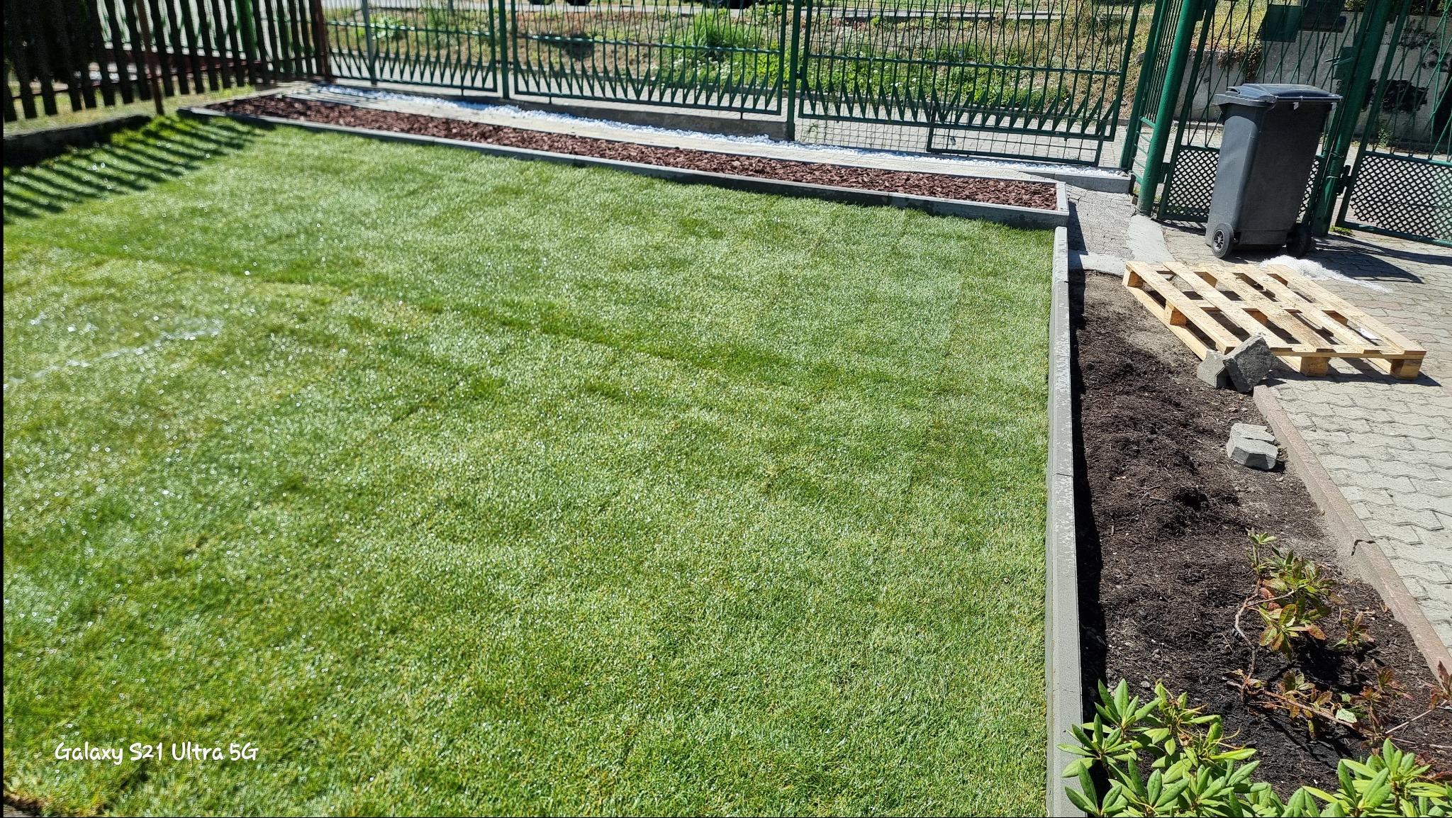 Świeżo ułożony trawnik z rolki, obrzeża z kostki, widoczny fragment rabaty z przygotowaną ziemią pod nasadzenia, paleta i kosz na śmieci w tle.