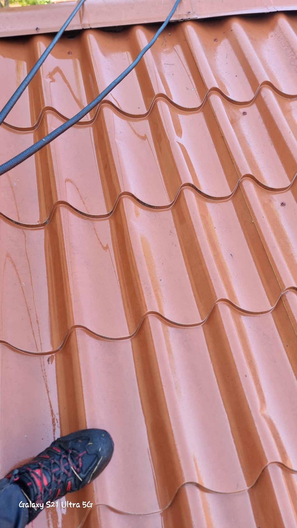 Błyszczące, mokre dachówki z blachy trapezowej w kolorze ceglastym, widziane z góry, z fragmentem buta robotnika w lewym dolnym rogu i wężem ogrodowym na górze.