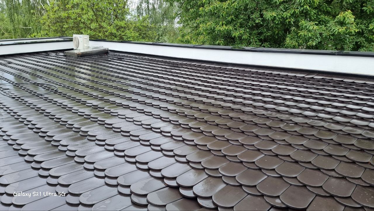 Dach pokryty ciemnymi, błyszczącymi dachówkami w kształcie łusek, z widocznym fragmentem klimatyzatora i zielenią w tle.