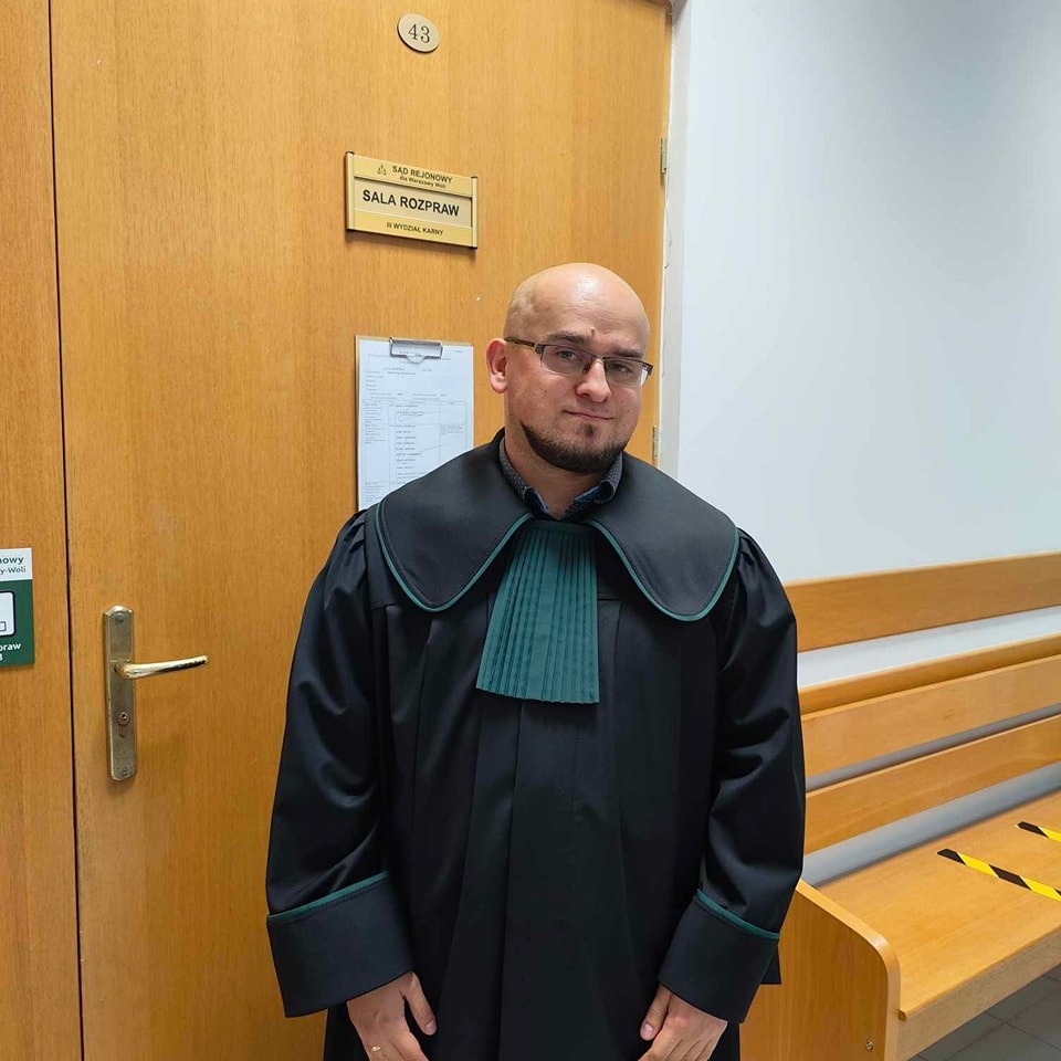 Portret mężczyzny w todze adwokackiej na tle drzwi z napisem 'Sala Rozpraw w Wydziale Karnym' i numerem 43, stojącego w korytarzu sądu.