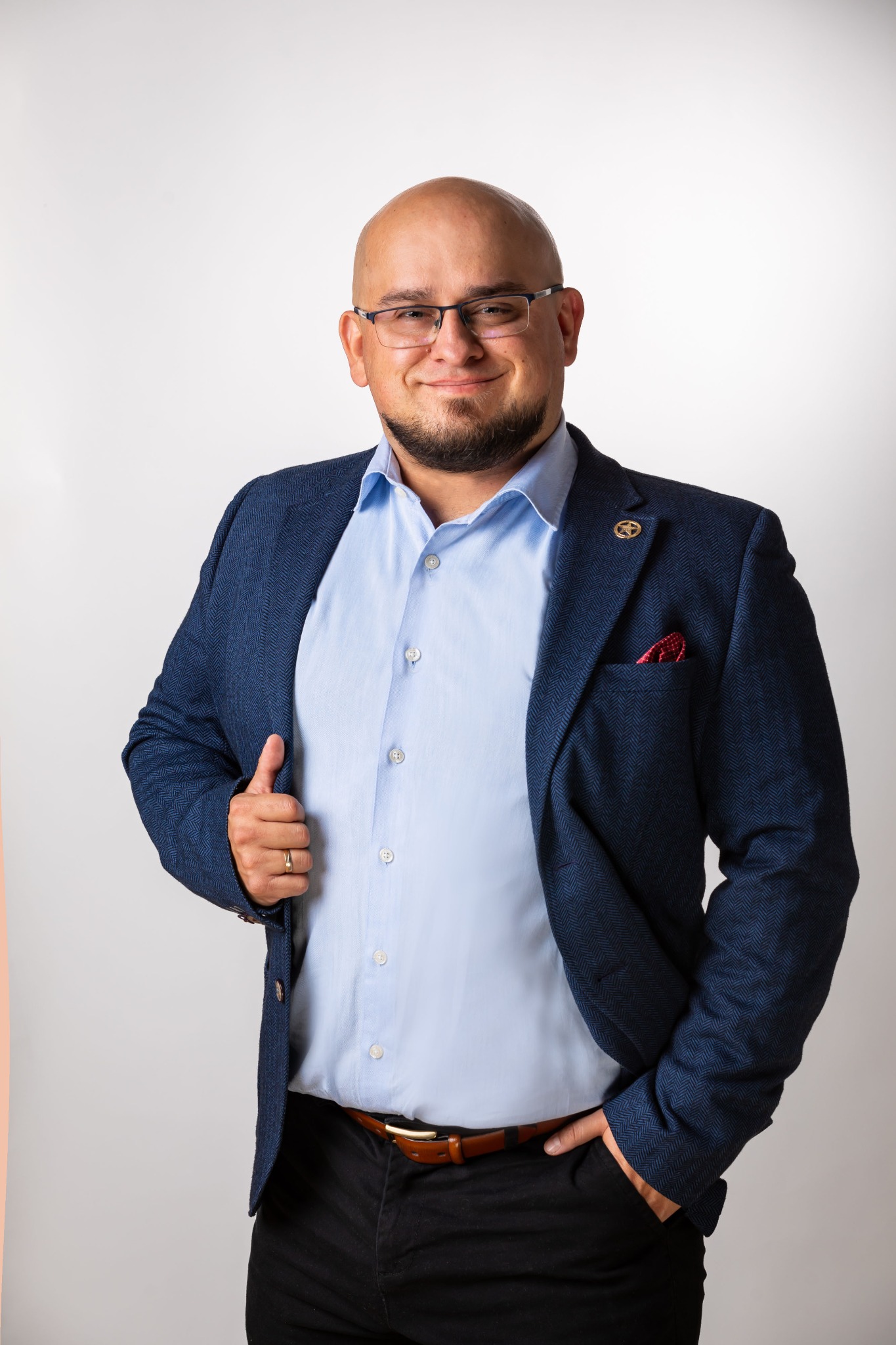 Portret uśmiechniętego mężczyzny w okularach, niebieskiej koszuli i marynarce w jodełkę, z chusteczką w kieszonce i złotą przypinką, pozującego na jasnym tle.