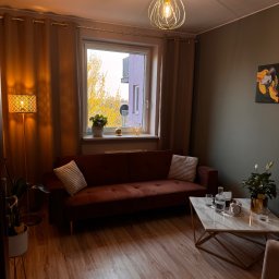 Pracownia Psychoterapii AGATA NIEDŹWIEDŹ-DĄBKOWSKA - Przytulny salon z brązową sofą, marmurowym stolikiem i lampą podłogową. Za oknem widok na jesienne drzewa i budynek. Ciepłe oświetlenie tworzy relaksującą atmosferę.