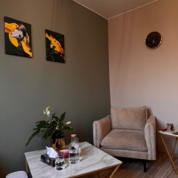 Pracownia Psychoterapii AGATA NIEDŹWIEDŹ-DĄBKOWSKA - Stylowy gabinet z beżowym fotelem, marmurowym stolikiem i złotymi akcentami. Na ścianie wiszą obrazy i zegar. Przytulne wnętrze, idealne do sesji terapeutycznej.