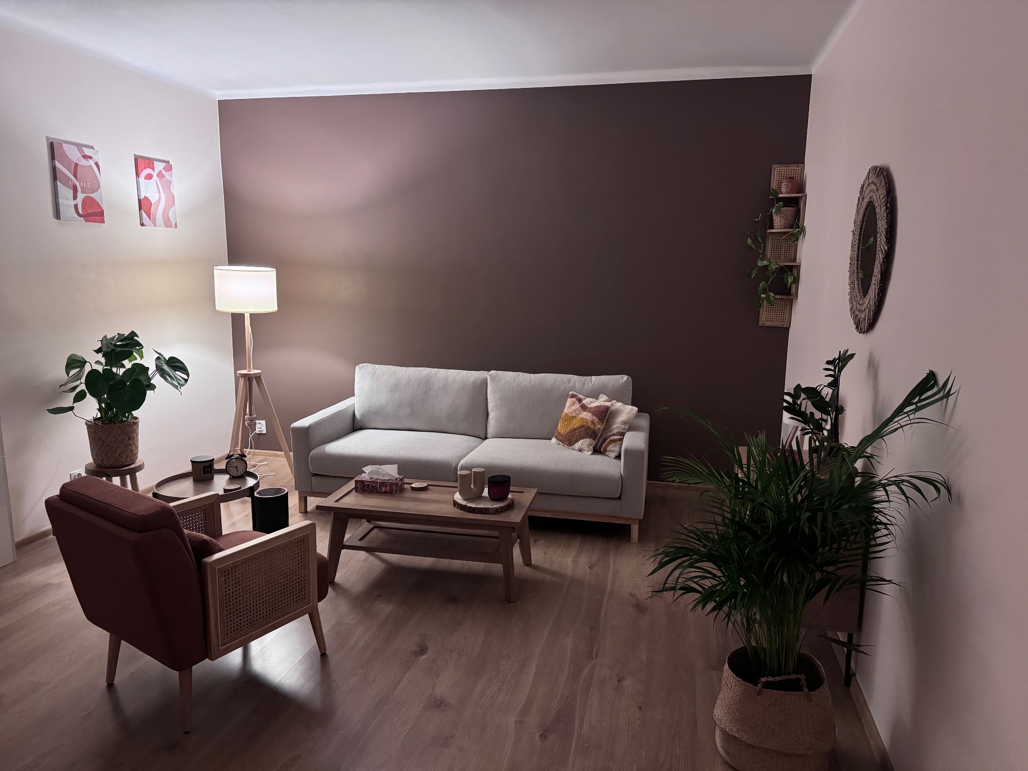 Ustawne wnętrze gabinetu psychoterapeuty z szarą sofą, drewnianym stolikiem kawowym, fotelem z elementami rattanu, roślinami doniczkowymi i lampą stojącą, utrzymane w stonowanej kolorystyce.