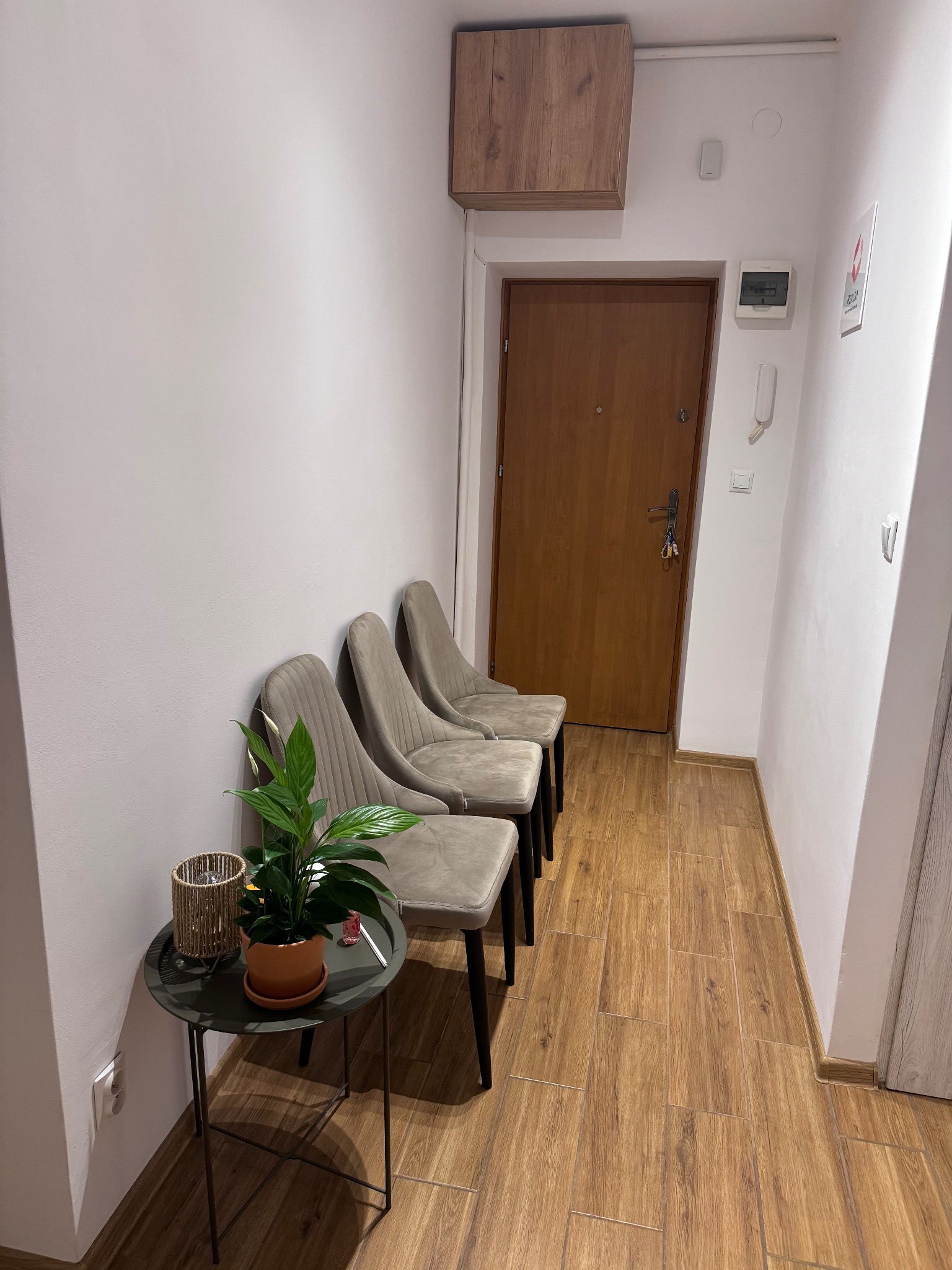 Poczekalnia z trzema krzesłami w odcieniach beżu, stolik z rośliną doniczkową i ozdobnym lampionem, drewniane panele podłogowe, brązowe drzwi wejściowe.
