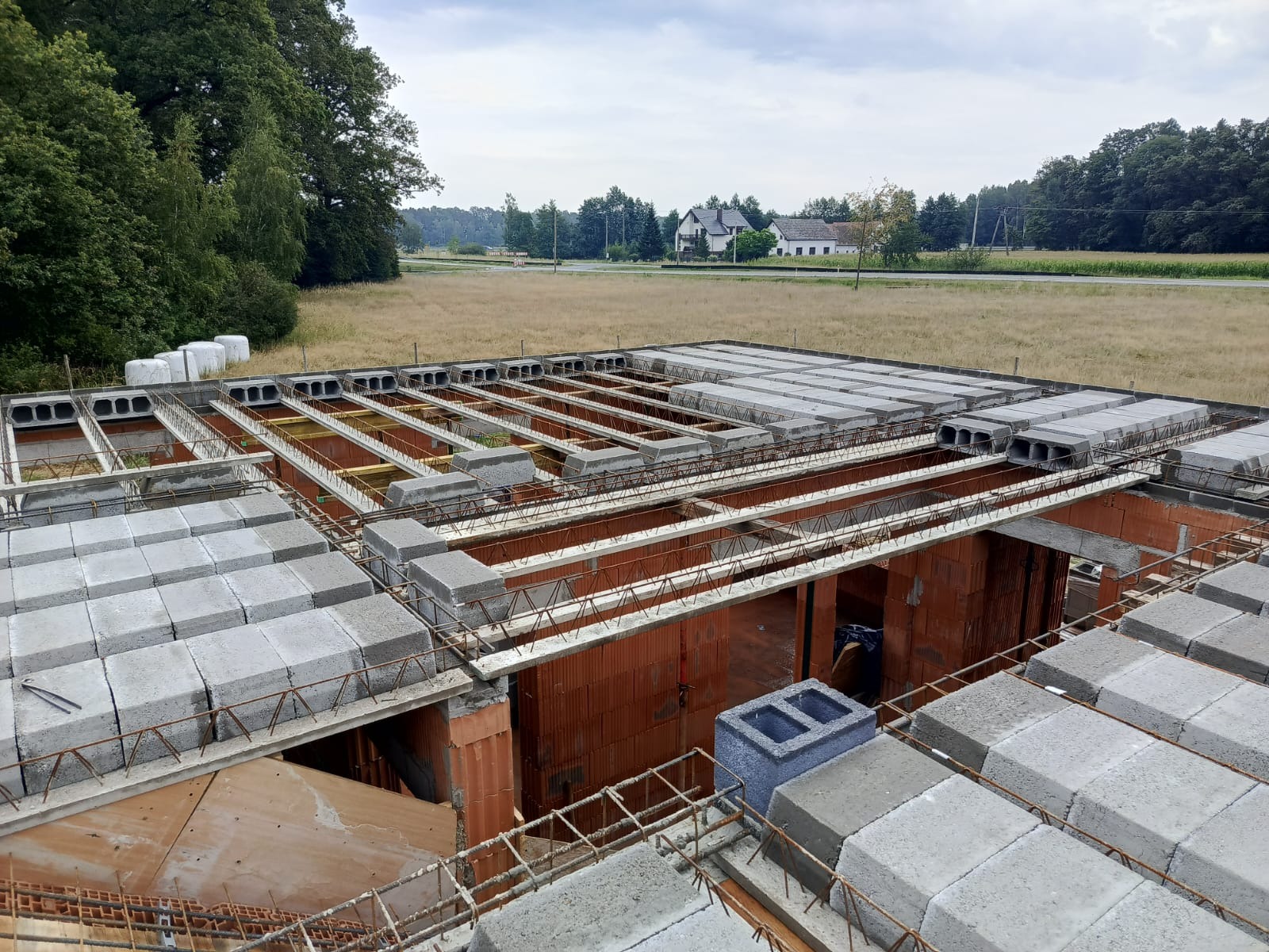 Wznoszenie stropu Teriva na budynku z czerwonej cegły, widok z góry na plac budowy z elementami szalunków i zbrojenia, w tle łąka i zabudowania.