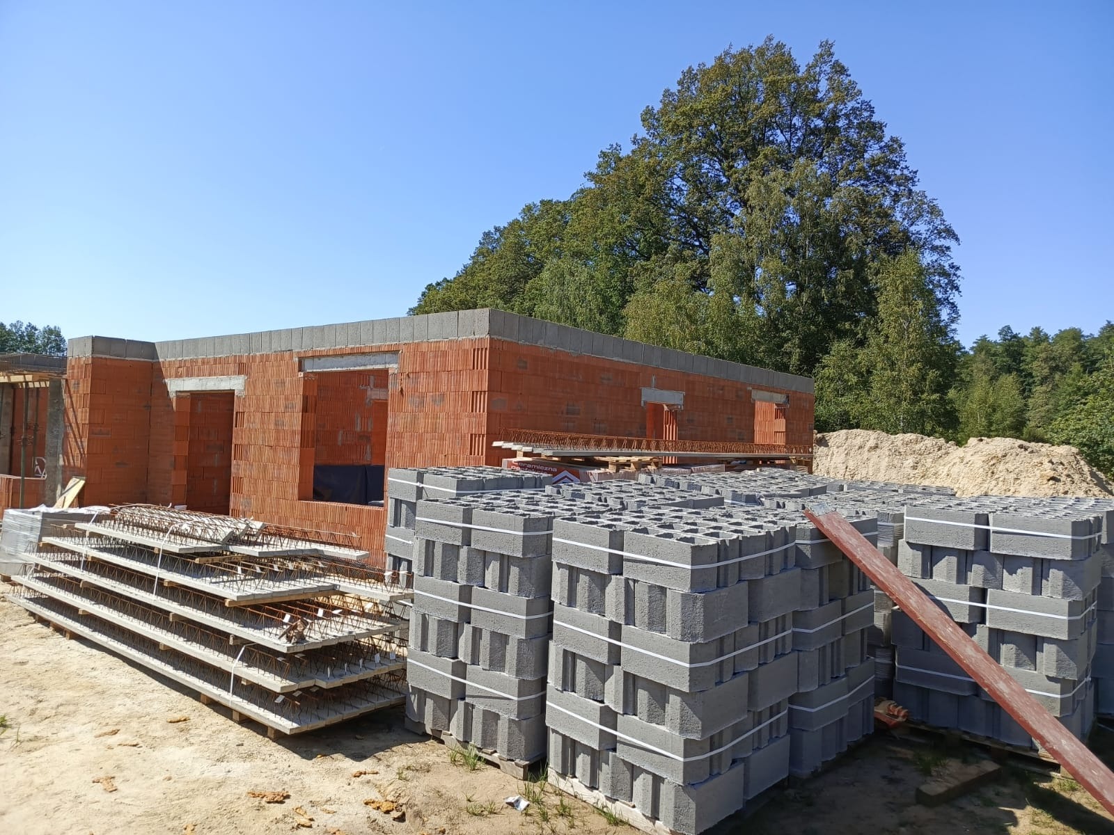 Wznoszone ściany budynku z czerwonej cegły ceramicznej, widoczne szare bloczki betonowe na paletach, zbrojenie stropu i drzewa w tle pod błękitnym niebem.