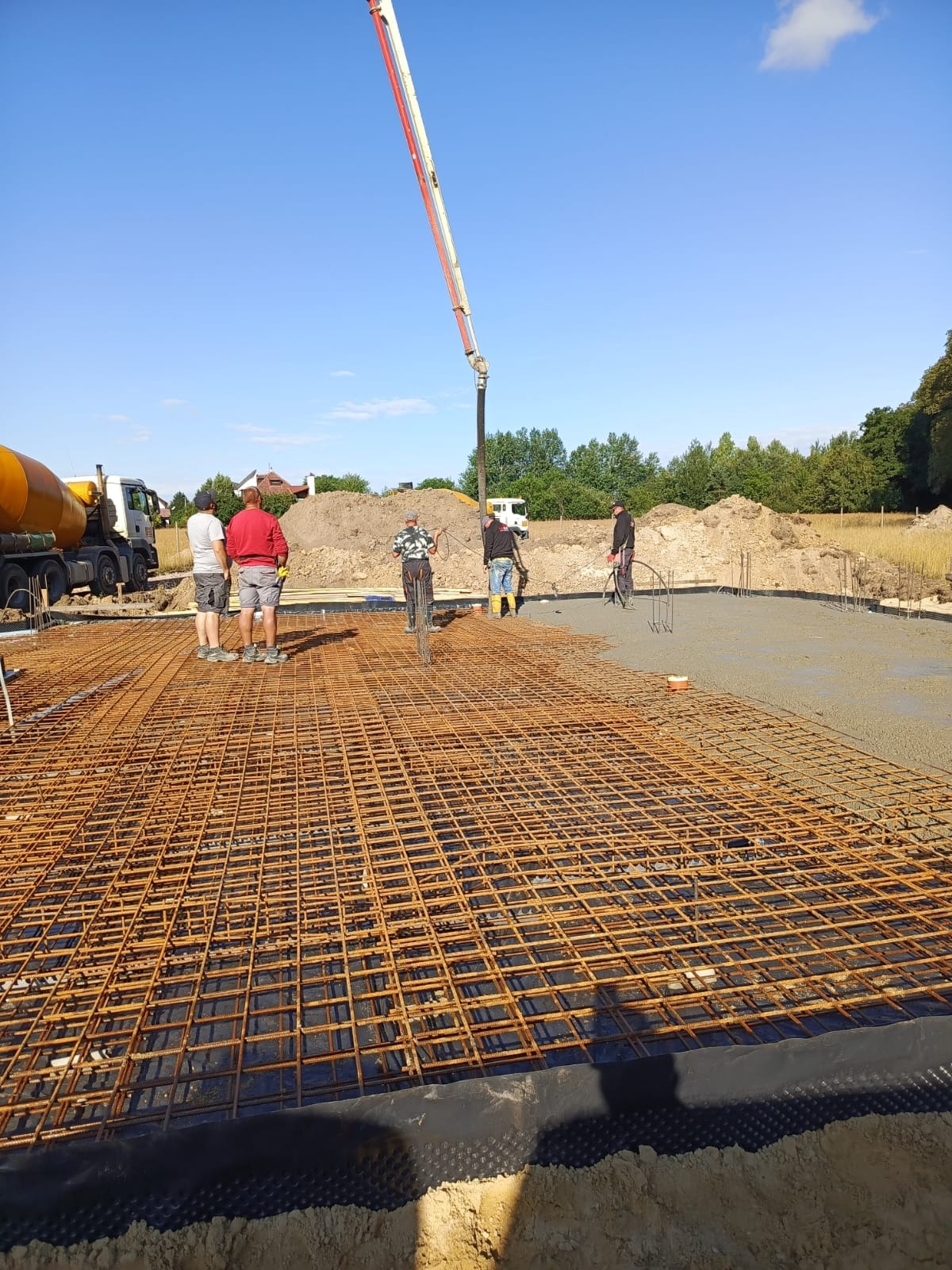 Wylewanie betonu na przygotowane zbrojenie fundamentu pod budowę, widoczny żółty betonomieszarka i wysięgnik pompy do betonu oraz grupa robotników w trakcie pracy.