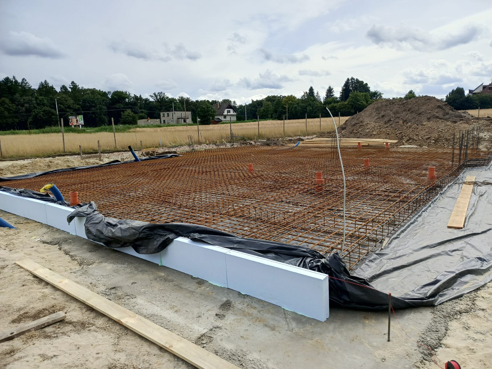 Zbrojenie płyty fundamentowej z widocznym szalunkiem z białego styropianu i instalacjami pionowymi, przygotowane do zalania betonem, w tle widoczny krajobraz.