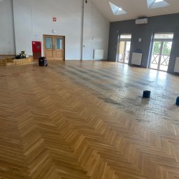 FIRMA USŁUGOWA PARKIETY Rafał Michalski - Duża sala z drewnianą podłogą ułożoną w jodełkę, częściowo odnowioną, widoczne ślady po szlifowaniu, w tle sprzęt do obróbki podłóg oraz elementy wyposażenia sali.