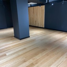 FIRMA USŁUGOWA PARKIETY Rafał Michalski - Jasna, drewniana podłoga z panelami ułożonymi ukośnie w przestronnym, nowoczesnym wnętrzu z ciemnymi ścianami i kolumną. Widoczne oświetlenie sufitowe.