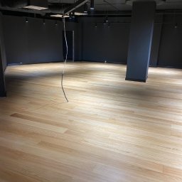 FIRMA USŁUGOWA PARKIETY Rafał Michalski - Jasne panele podłogowe w nowym pomieszczeniu z szarą ścianą i surowym oświetleniem, widoczne wiszące przewody.