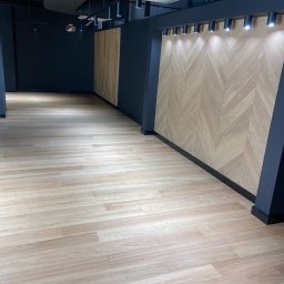 FIRMA USŁUGOWA PARKIETY Rafał Michalski - Jasna podłoga z paneli w Kamieńcu, ściana wykończona drewnianą mozaiką w jodełkę, sufit z czarnymi lampami punktowymi.