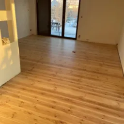 Jasne, drewniane deski podłogowe z widocznymi słojami, ułożone w nowym pokoju z dużymi drzwiami balkonowymi i widokiem na zimowy krajobraz.