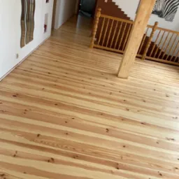 Jasna, drewniana podłoga z widocznymi słojami, świeżo po renowacji, z drewnianą balustradą schodów i lustrem na ścianie w tle.