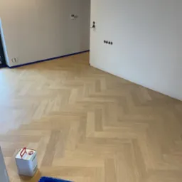 Jasna, nowa podłoga z parkietu ułożonego w jodełkę francuską, zabezpieczona niebieską taśmą malarską przy ścianach; obok puszka lakieru i kuweta z pędzlem.
