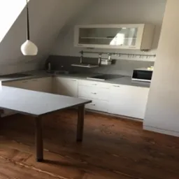 Kuchnia w skosach poddasza z białymi szafkami, blatem w odcieniach szarości i drewnianą podłogą z wyraźnym usłojeniem, oświetlona wiszącymi lampami.