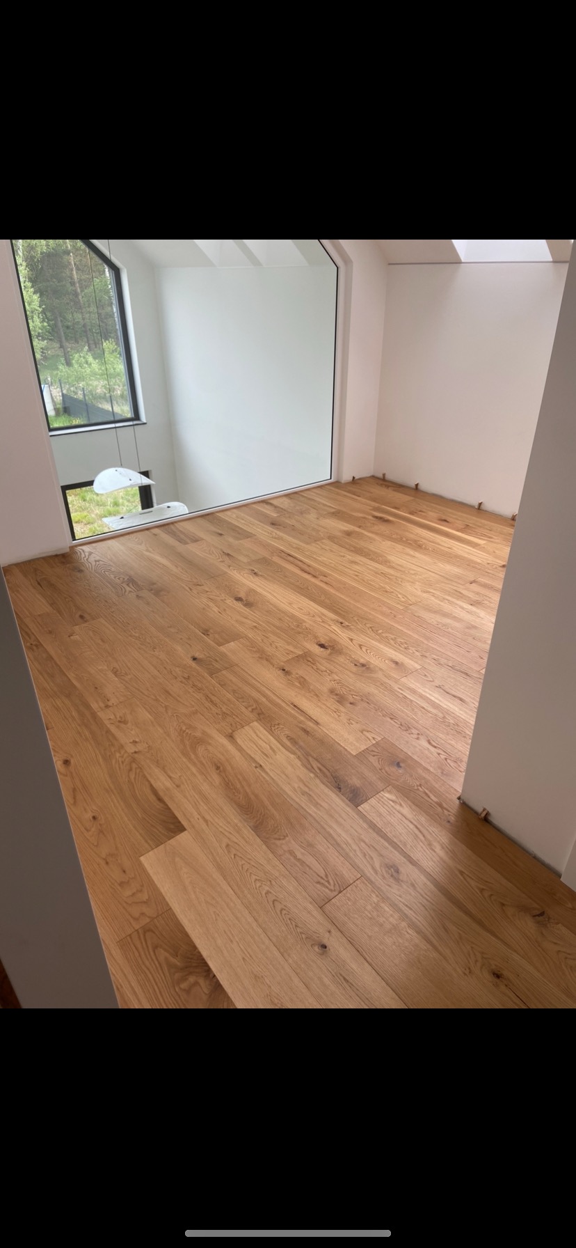 Jasna, dębowa podłoga ułożona w pustym, nowoczesnym pokoju z dużym oknem i białymi ścianami, widoczne kliny dystansowe przy ścianie.