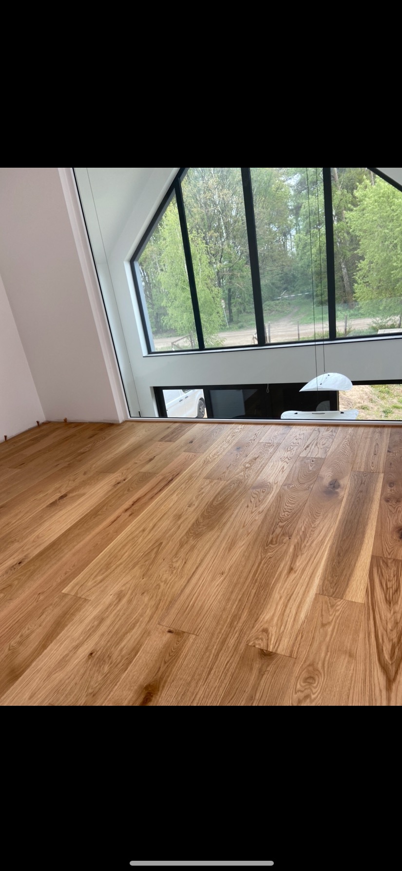 Jasny parkiet dębowy ułożony w nowoczesnym wnętrzu z dużym oknem trapezowym z widokiem na zieleń; listwy przypodłogowe jeszcze nie zamontowane.